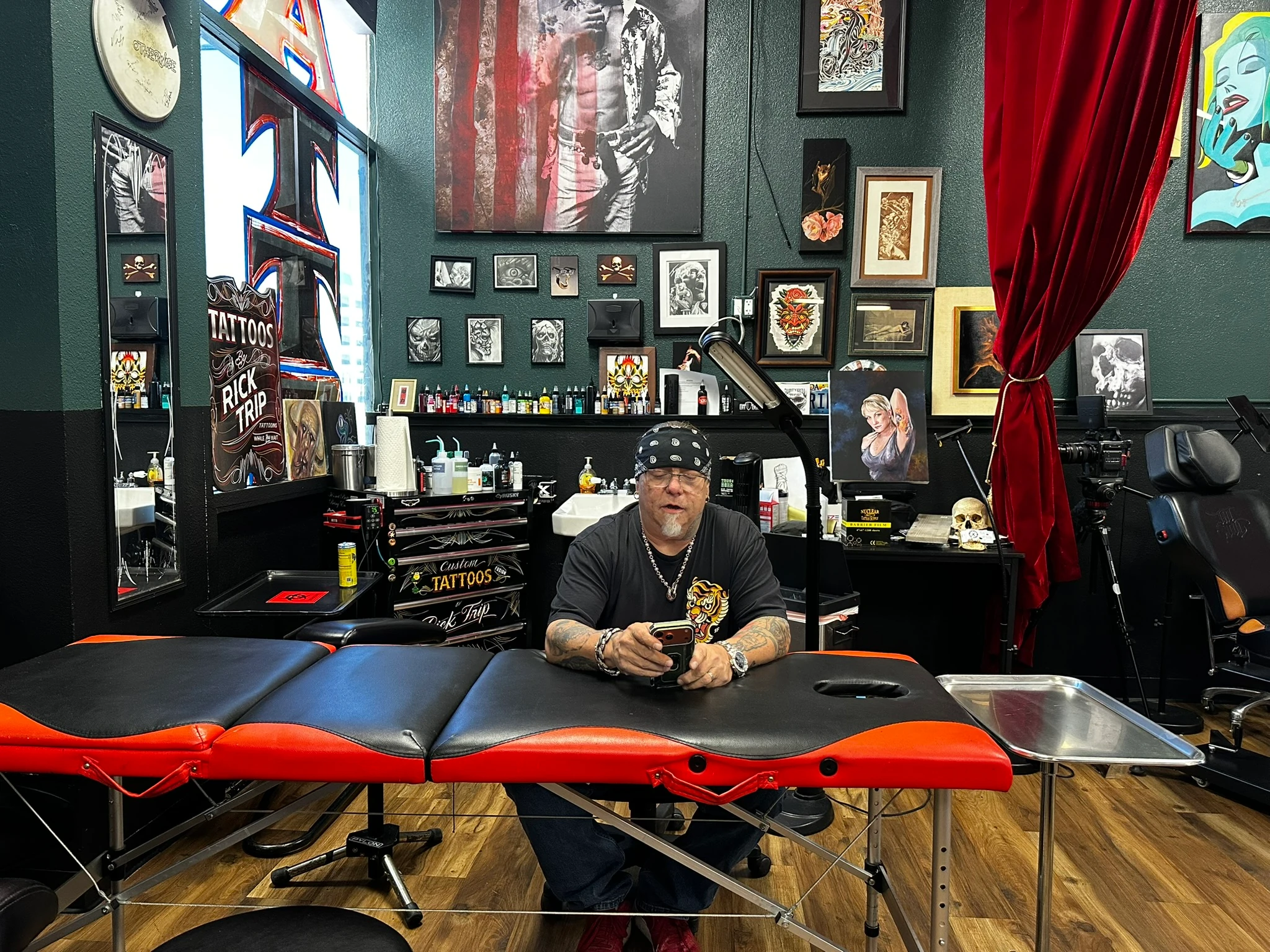 Rick Trip seated behind a red-and-black tattoo table in his studio, surrounded by framed artwork and tattoo equipment