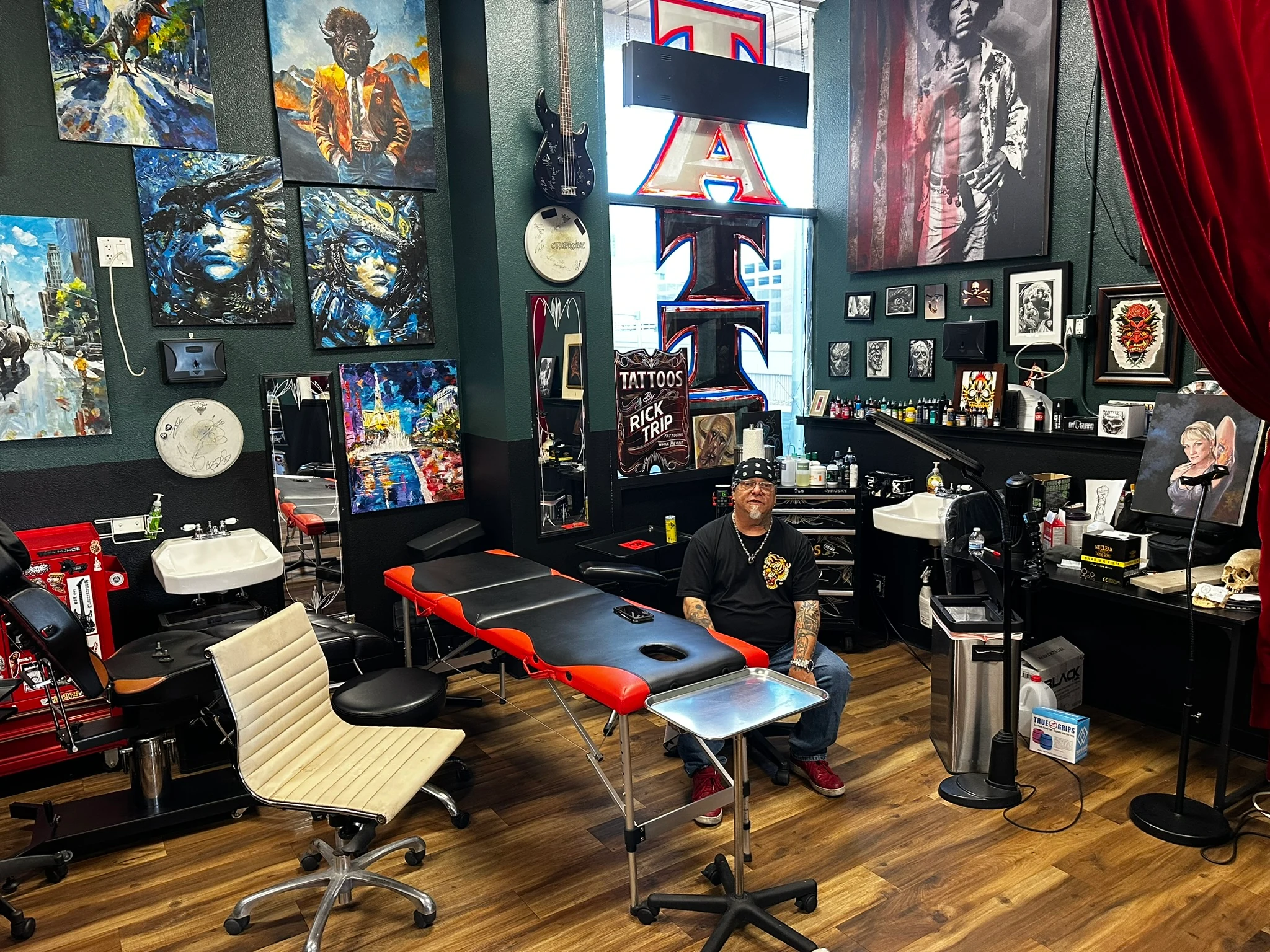 Rick Trip facing the camera in his tattoo studio, with framed artwork, red curtains and black-and-red tattoo furniture