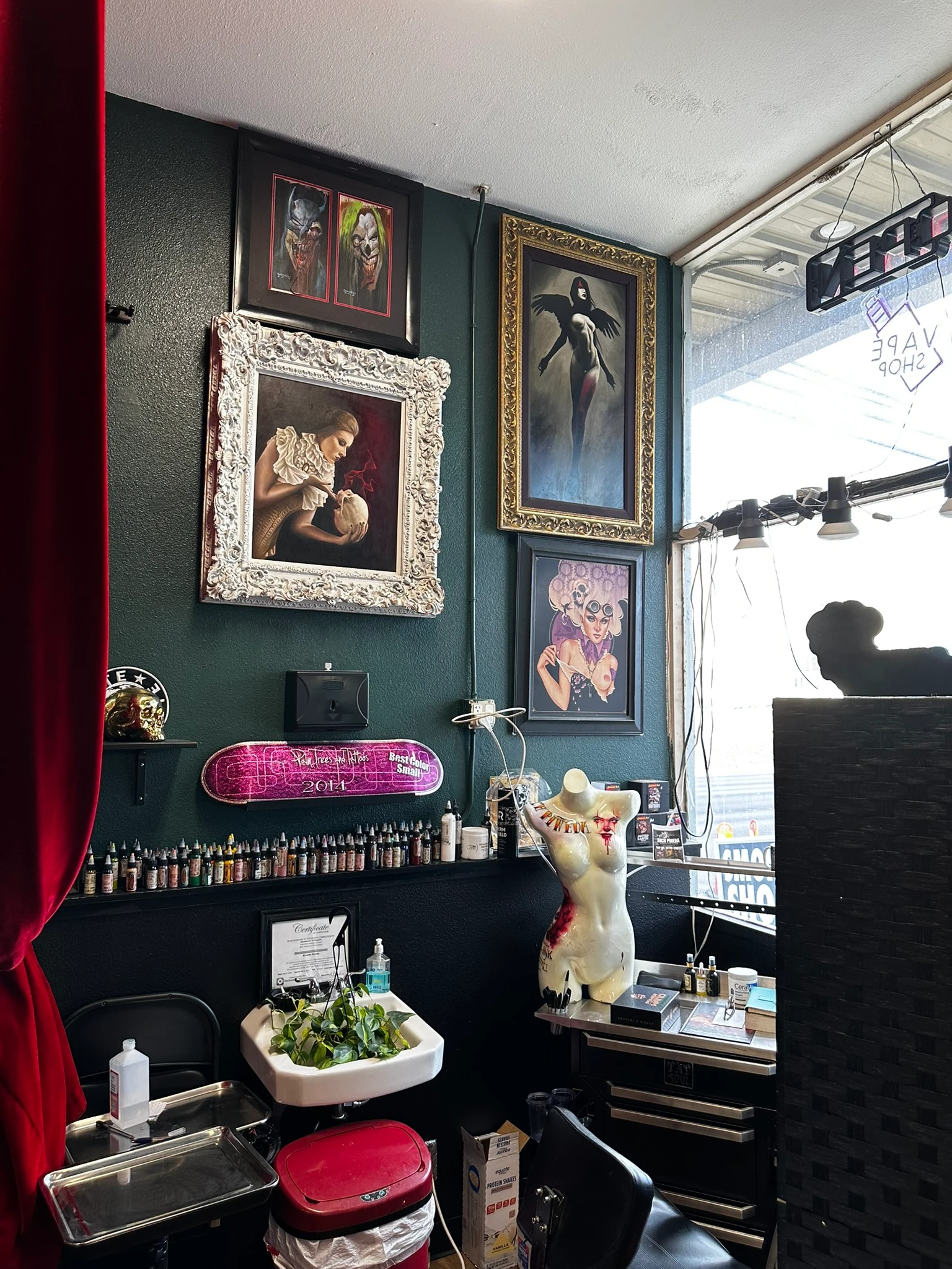 Close view of framed artwork, tattoo ink bottles, a sink and studio objects beside a window and red curtain